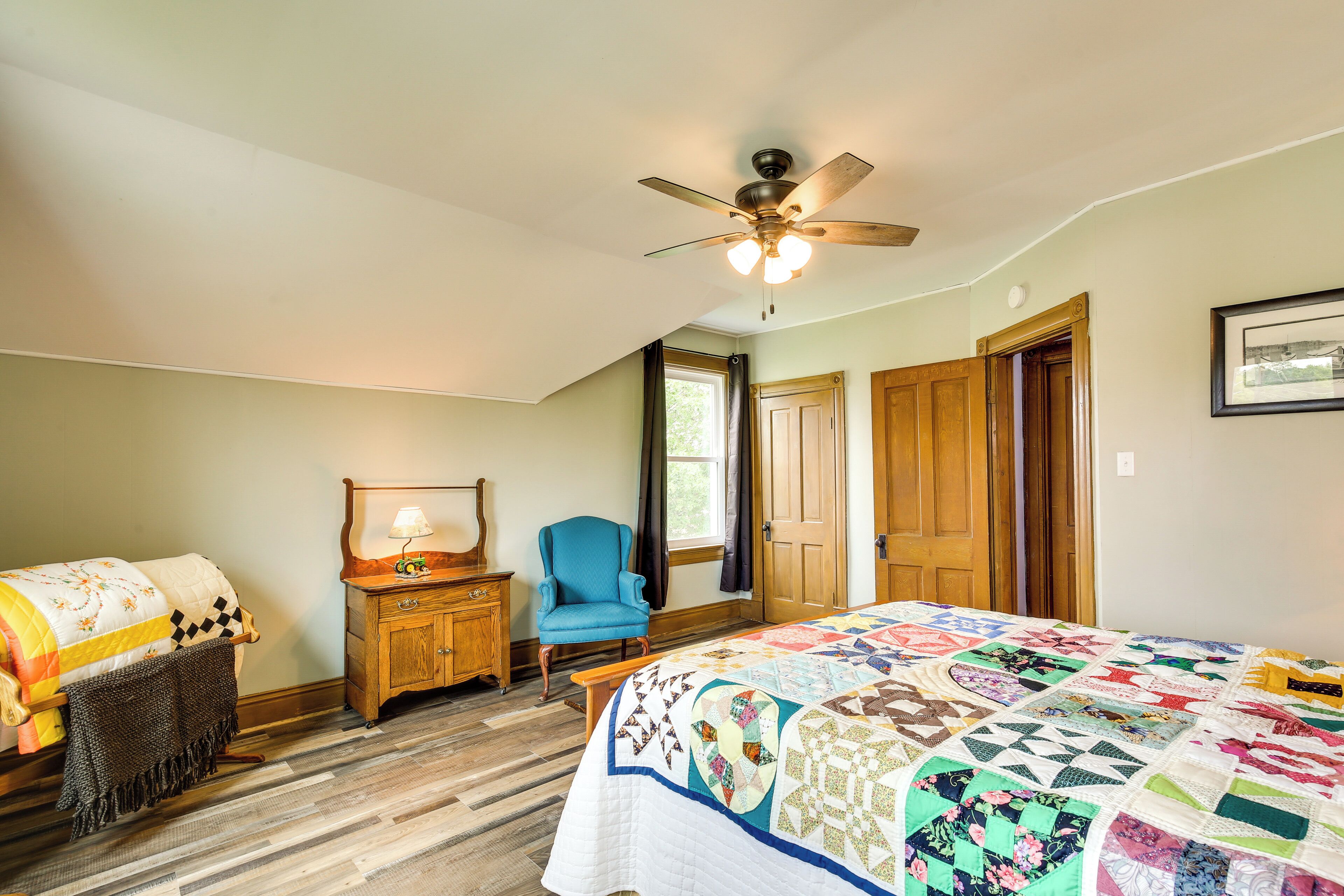 Une chambre à coucher avec un grand lit, un ventilateur de plafond, une commode en bois, un fauteuil bleu et une fenêtre avec des rideaux.