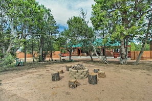 A rustic outdoor area with a stone fire pit surrounded by tree stumps, a wooden bench, and a small table. - Fire Pit, Fenced Yard: Overgaard Cabin Near Hiking (Overgaard)