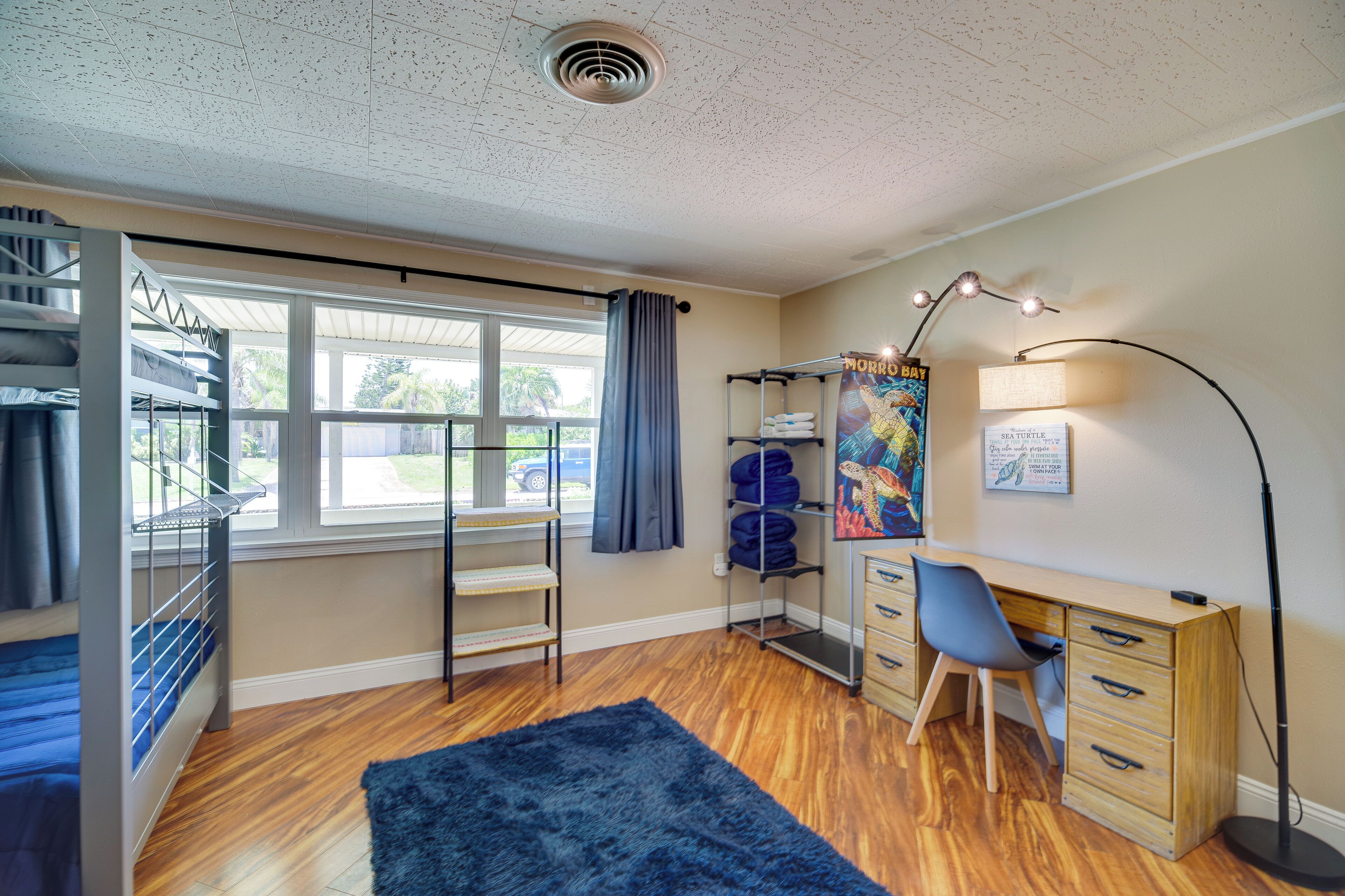 A dormitory room with bunk beds, a desk, a chair, a lamp, and a large window.