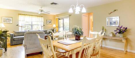 A dining area with a wooden table and white chairs, a ceiling fan, and a window with curtains.