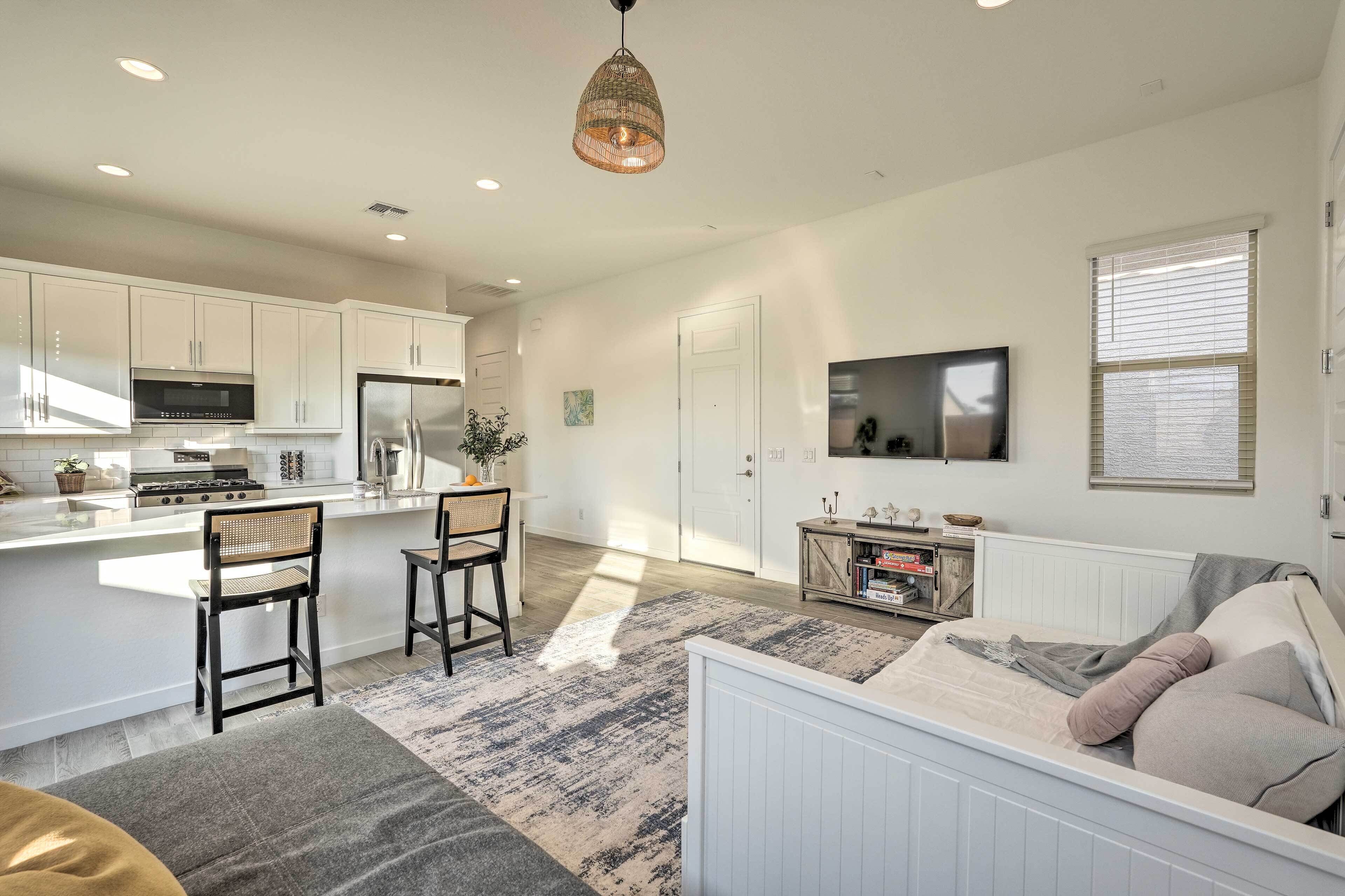 A modern kitchen with white cabinets, a built-in stove, and a large island. There is a dining area with two chairs and a table, a living room with a sofa and a TV, and a large window with blinds.
