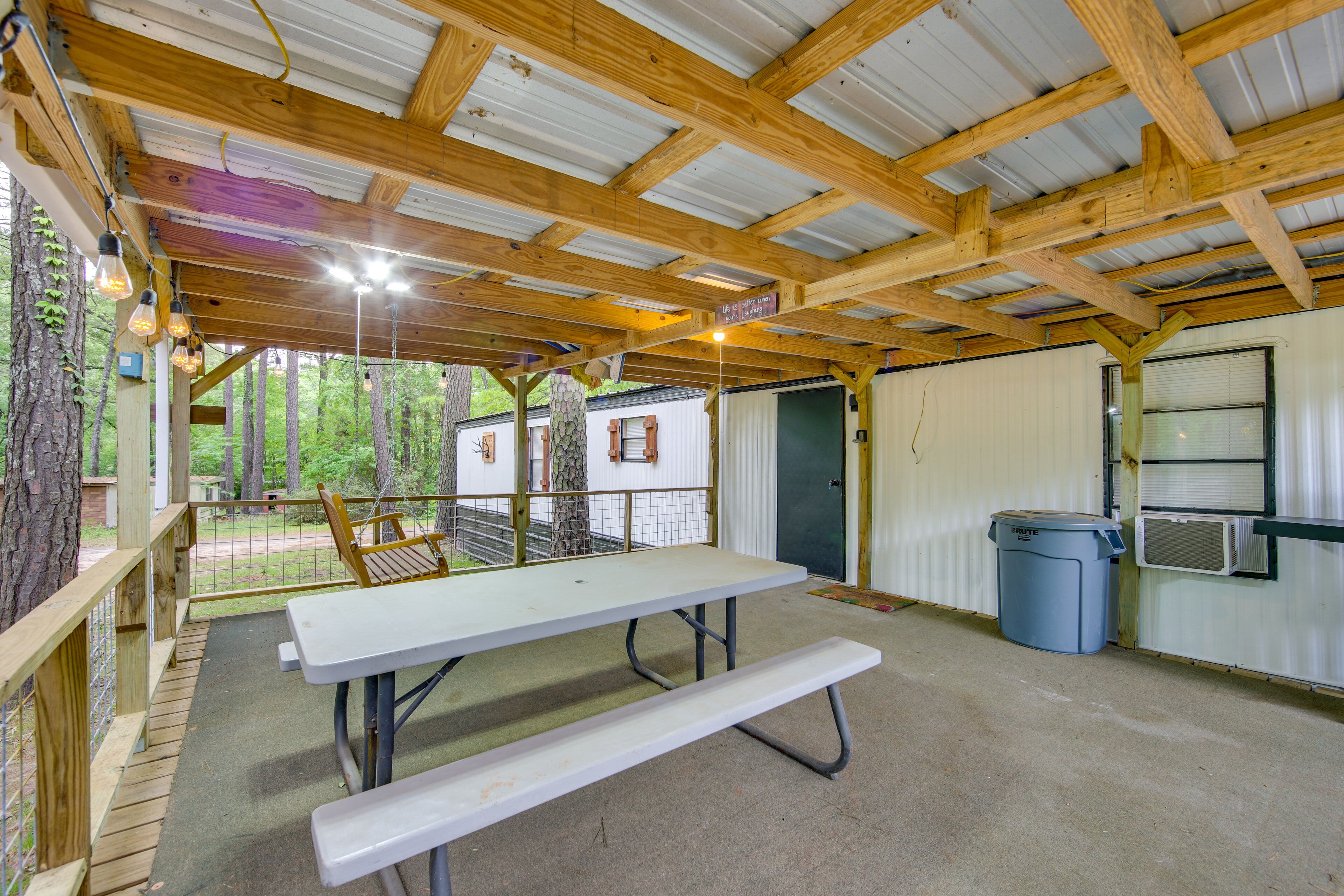 A covered outdoor area with a picnic table, a trash bin, and a view of the surrounding trees.