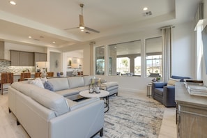 A spacious living room with a large sectional sofa, a coffee table, and a seating area with a blue sofa. There is a kitchen in the background with modern appliances and a dining area with wicker chairs. - Luxury Spring Training Home Base in Goodyear! (Goodyear)