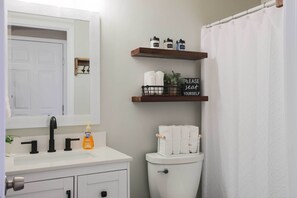 A bathroom with a white sink, black faucet, and a toilet. There are wooden shelves above the toilet holding toiletries and a sign that says "PLEASE SEAT YOURSELF". - Charming Anchorage Townhouse w/ Fire Pit! (Anchorage)