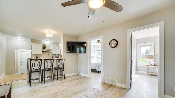 A spacious living area with a wooden floor, a ceiling fan, a flat-screen TV mounted on the wall, and a kitchen area with a bar counter and chairs. - Cozy Lewiston Home - Near Lakes & Restaurants! (Lewiston)