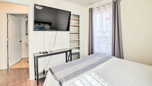 Ein Schlafzimmer mit einem Bett, einem an der Wand montierten Fernseher, einem Schreibtisch mit Stuhl und einem fenster mit Vorhängen.