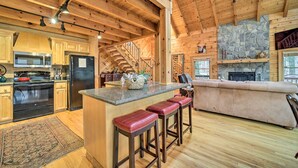 A spacious kitchen with wooden cabinets, a central island with stools, a stone fireplace, and a cozy seating area. - Walk to Golfing: Grand Maggie Valley Cabin! (Maggie Valley)