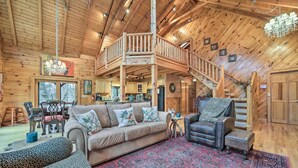 A spacious log cabin interior with a central living area, a dining area, and a kitchen. A wooden staircase connects the living area to an upper deck. - Walk to Golfing: Grand Maggie Valley Cabin! (Maggie Valley)