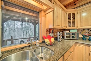 A kitchen with wooden cabinets, a granite countertop, a double sink, and a window overlooking a forest. - Walk to Golfing: Grand Maggie Valley Cabin! (Maggie Valley)