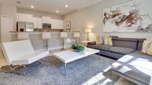 Ein modernes Wohnzimmer mit einem weißen Sofa, einem grauen Sofa und einem weißen Couchtisch. Es gibt eine Küchenzeile mit weißen Schränken und einer Mikrowelle. Ein Deckenventilator ist sichtbar, und ein großes abstraktes Gemälde hängt an der Wand.