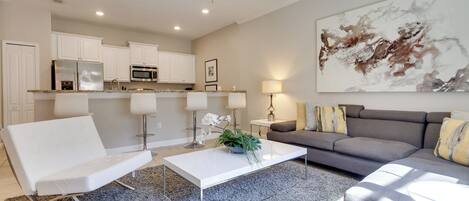 A modern living room with a white sofa, a grey sofa, and a white coffee table. There is a kitchen area with white cabinets and a microwave. A ceiling fan is visible, and a large abstract painting hangs on the wall.