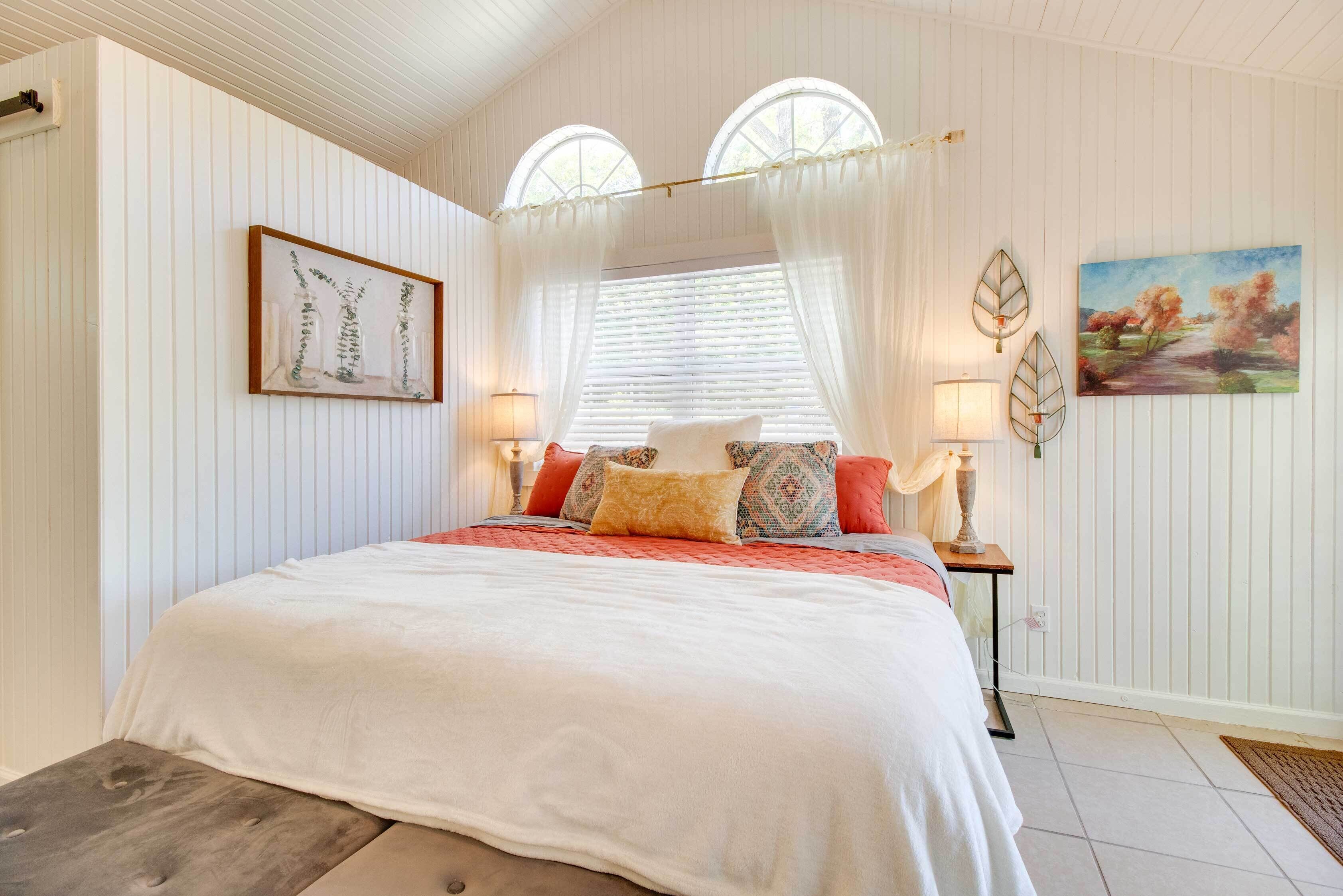 A bedroom with a large bed, two bedside lamps, a window with curtains, and a painting on the wall.