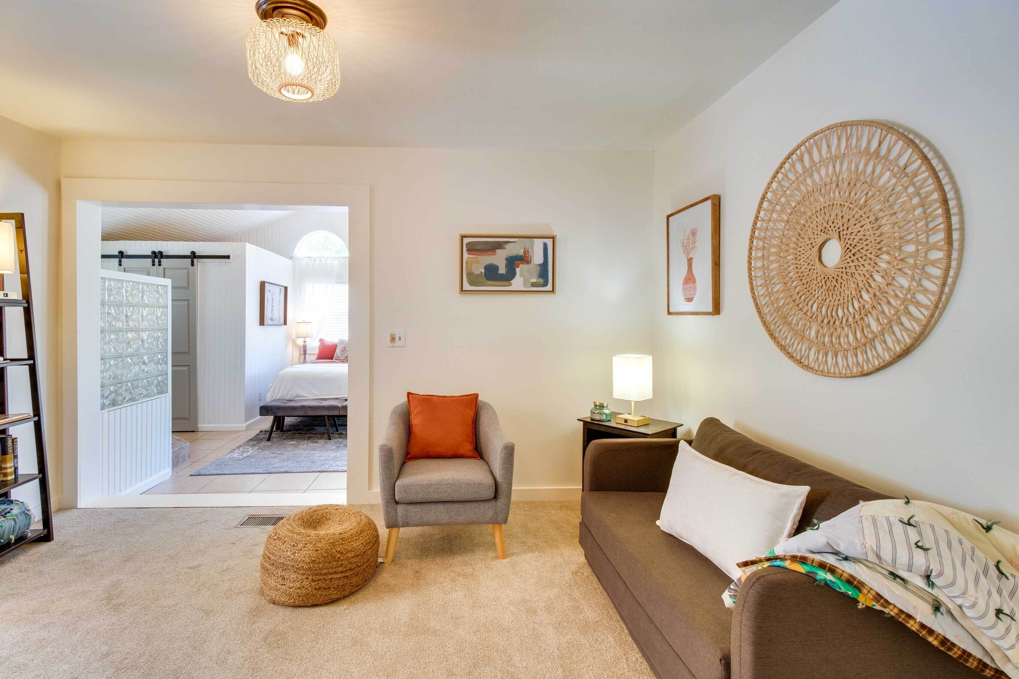 A cozy living room with a grey armchair, a brown sofa, and a woven rug. There is a door leading to another room, a wall-mounted lamp, and a framed picture.