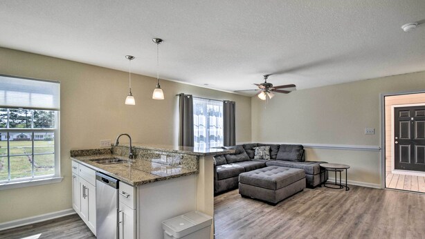 A modern kitchen with granite countertops, a dishwasher, and a window. There is a gray sectional sofa and a small table in the living area. - Cozy Cape Charles Apartment: Walk to Beaches! (Cape Charles)