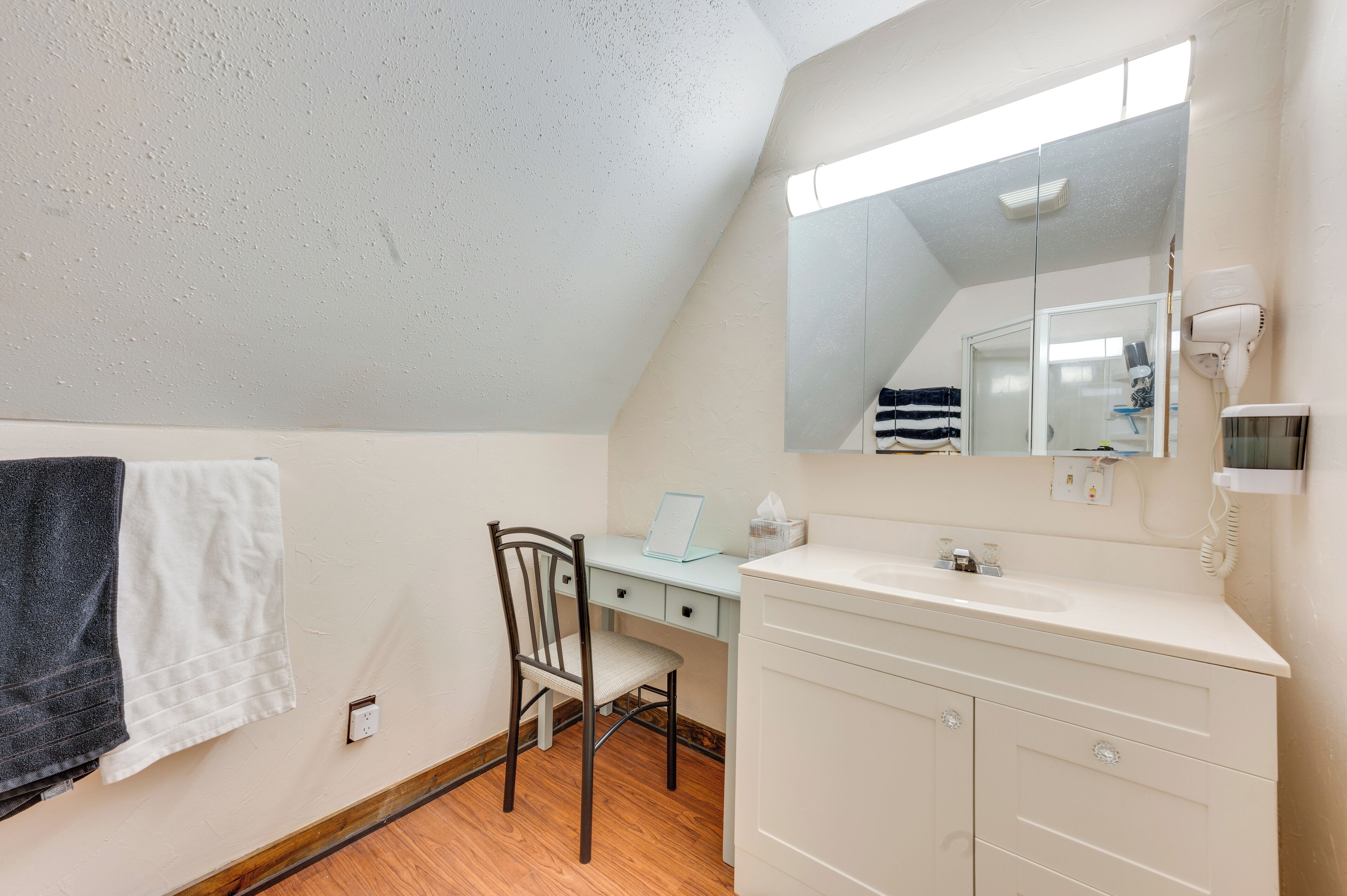 Una habitación pequeña y bien iluminada, con piso de madera, un escritorio con silla, un tocador blanco con espejo y una toalla colgada en la pared.
