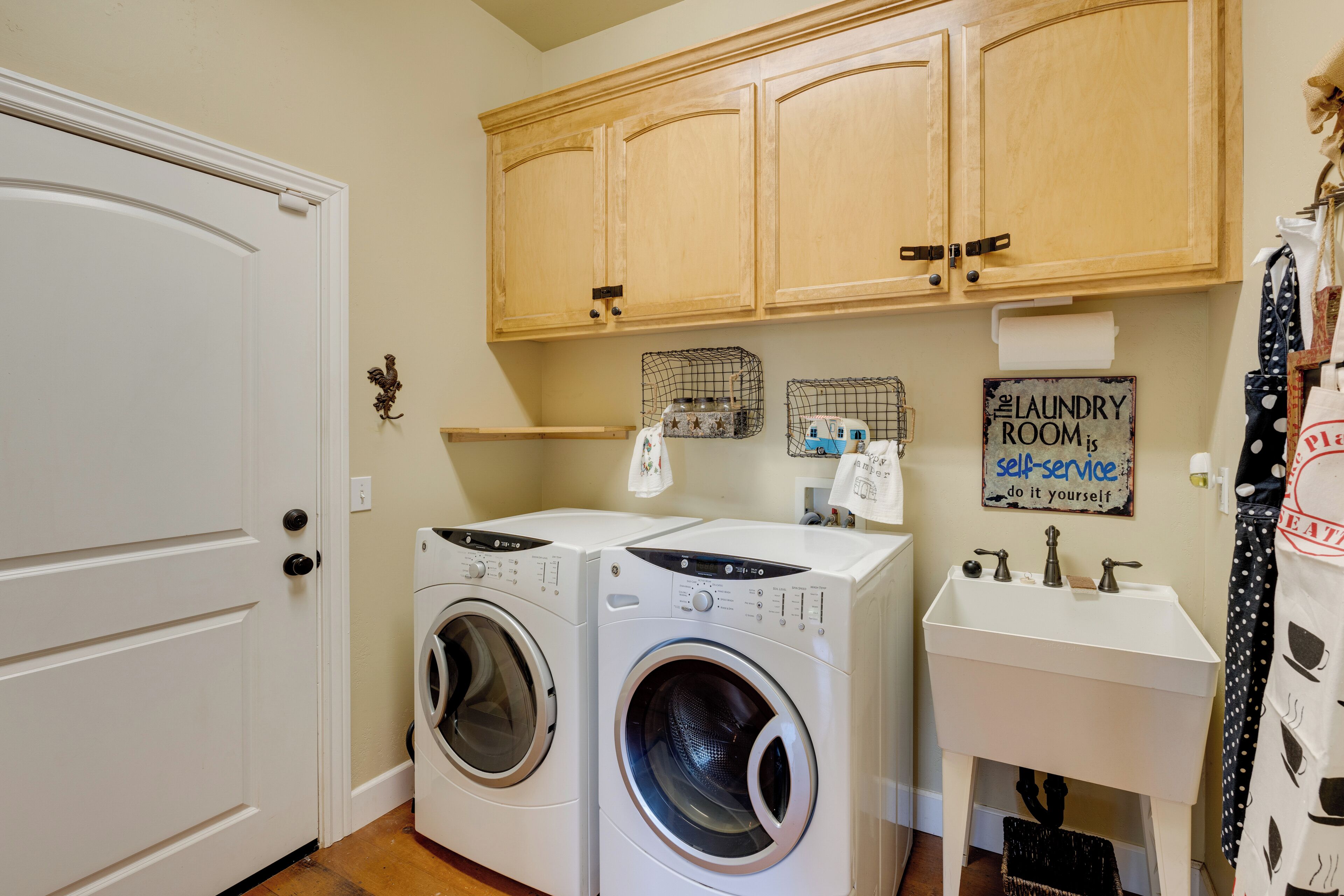 A laundry room with two front-loading washing machines, wooden cabinets above, a sink, and a sign that says "The Laundry Room is self-service."