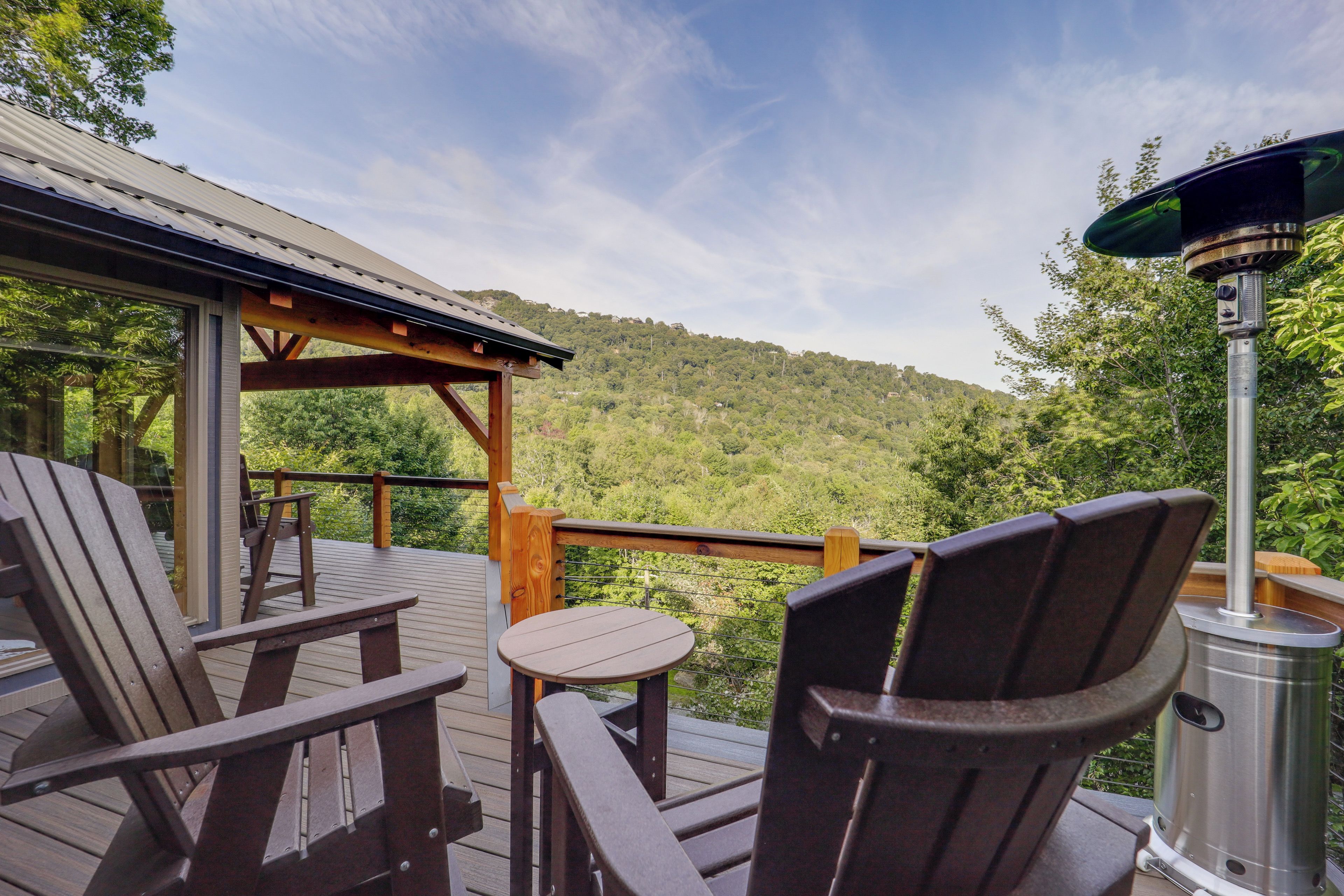 Eine Holzterrasse mit Gartenmöbeln, einem Metallgrill und Blick auf einen bewaldeten Hügel.