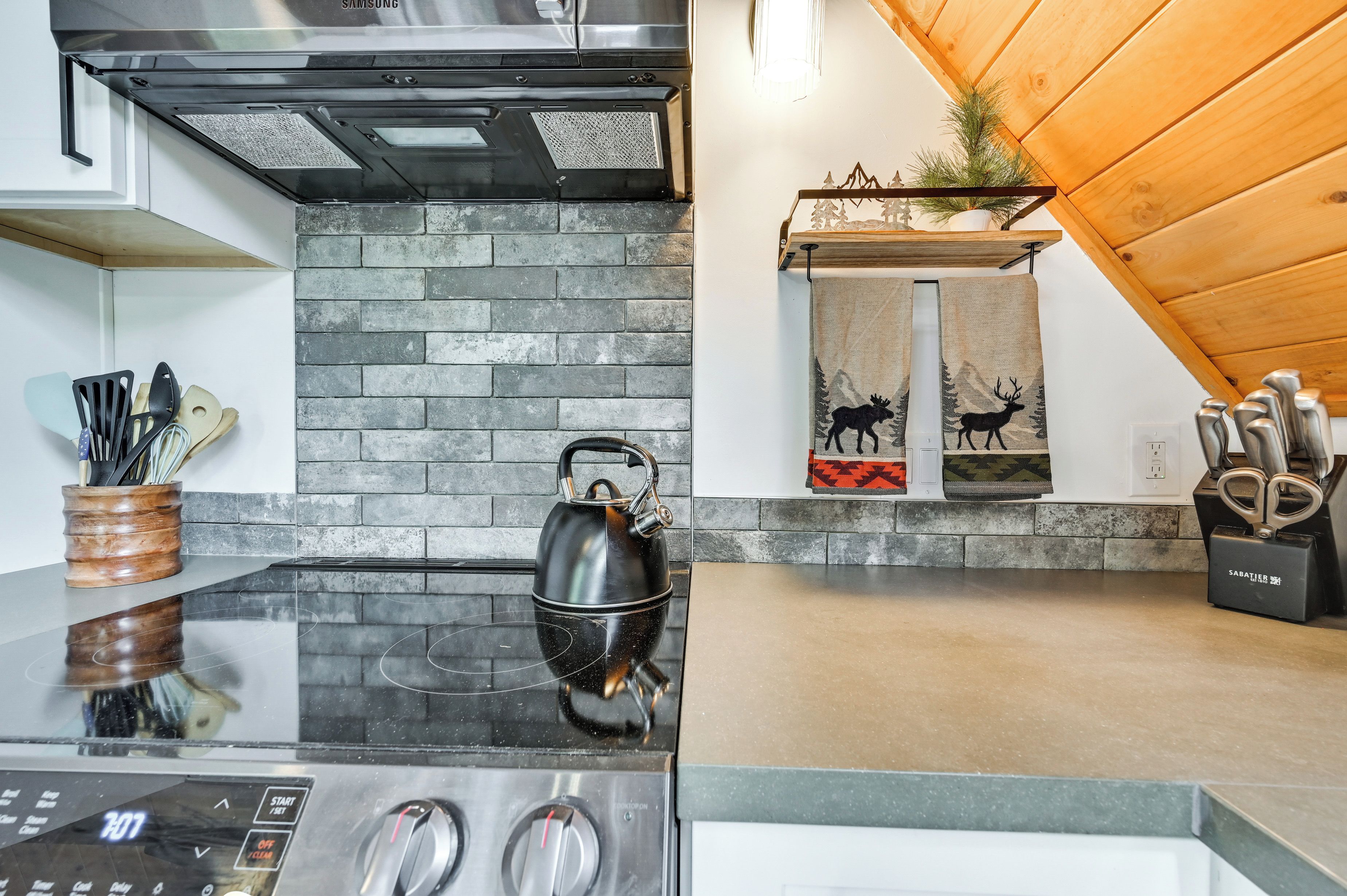 A modern kitchen with a black induction cooktop, a stainless steel range hood, and a brick backsplash.