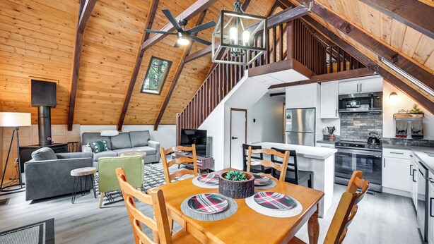 A spacious loft with a high ceiling, exposed wooden beams, a dining area with a wooden table and chairs, a kitchen with modern appliances, and a cozy living area with a sofa and a fireplace. - Deck & Grill: A-frame Cabin in Munds Park! (Munds Park)