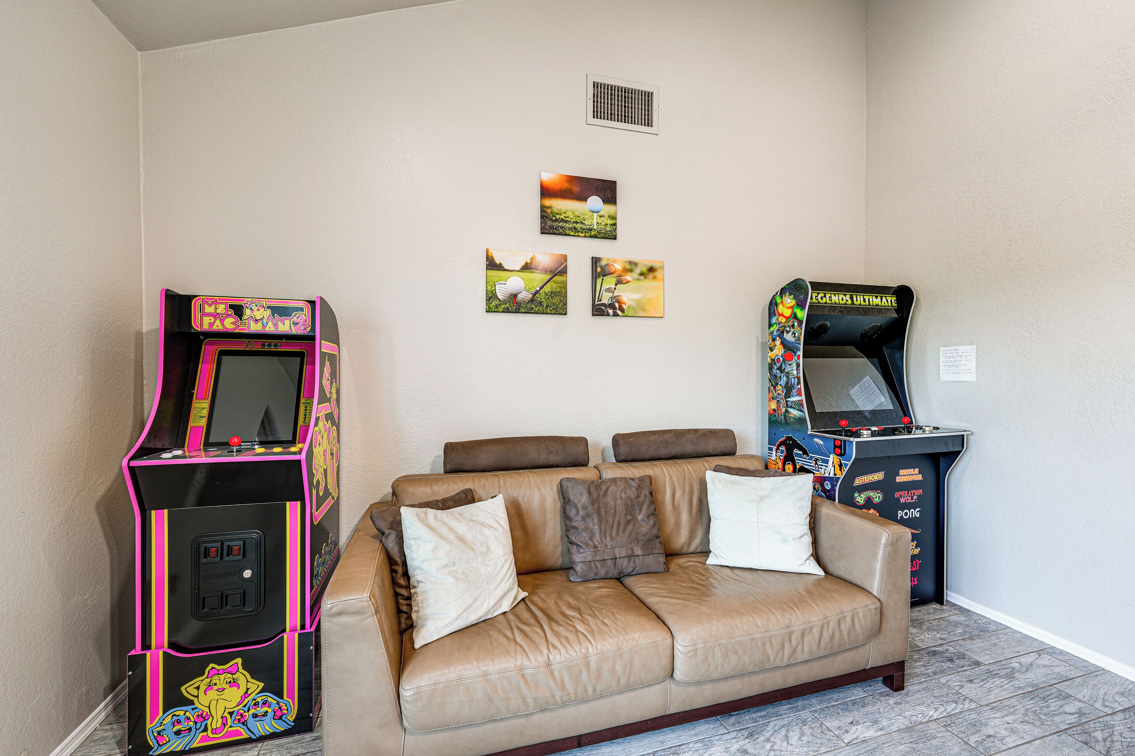A room with a beige leather couch, two arcade machines, and three framed pictures on the wall.