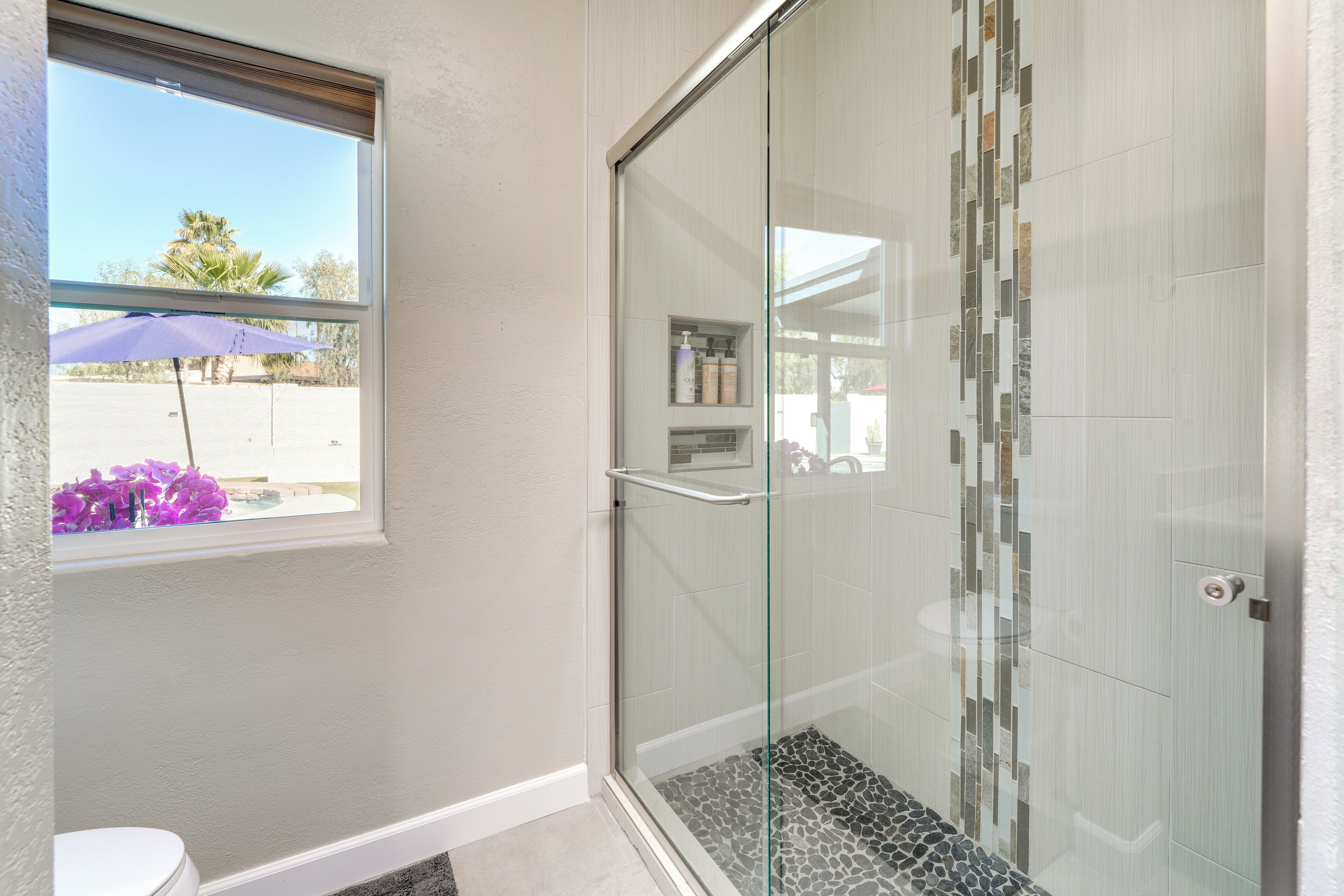 A bathroom with a glass shower enclosure, a toilet, and a window with a view of the outside.