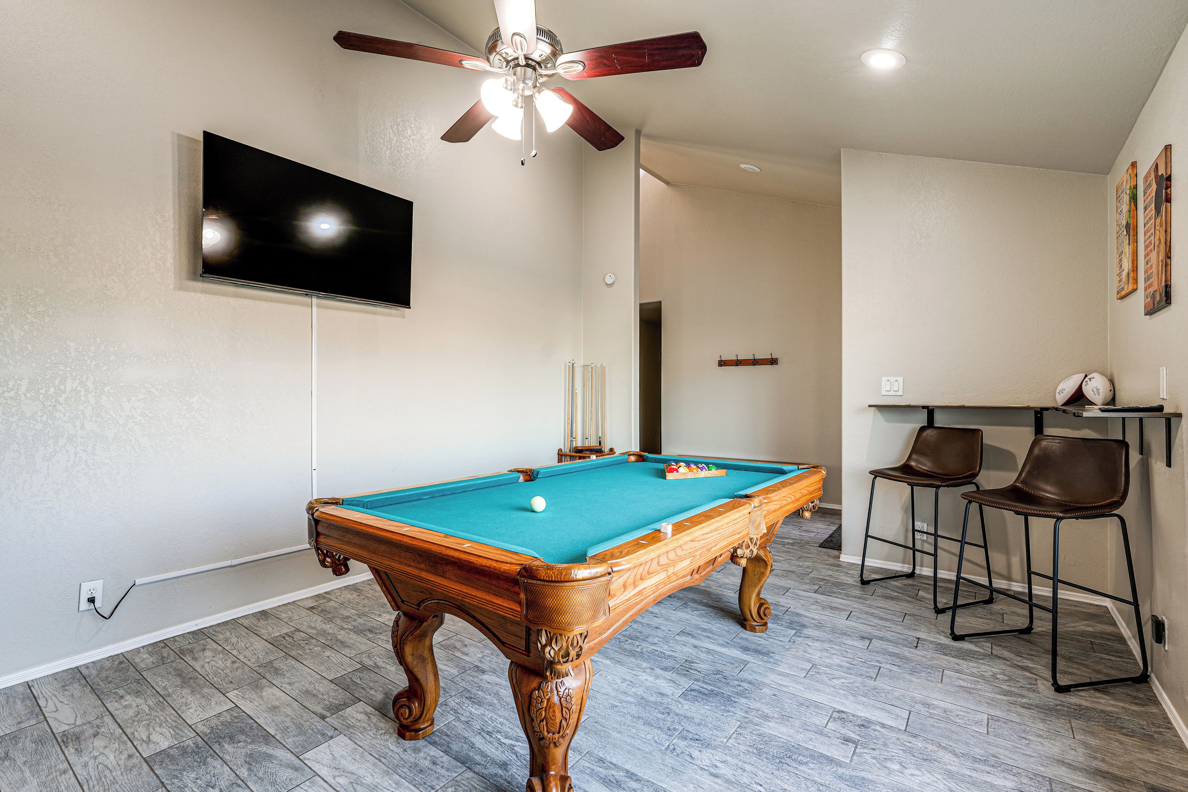 Une table de billard, un ventilateur de plafond, un téléviseur fixé au mur et deux tabourets de bar.