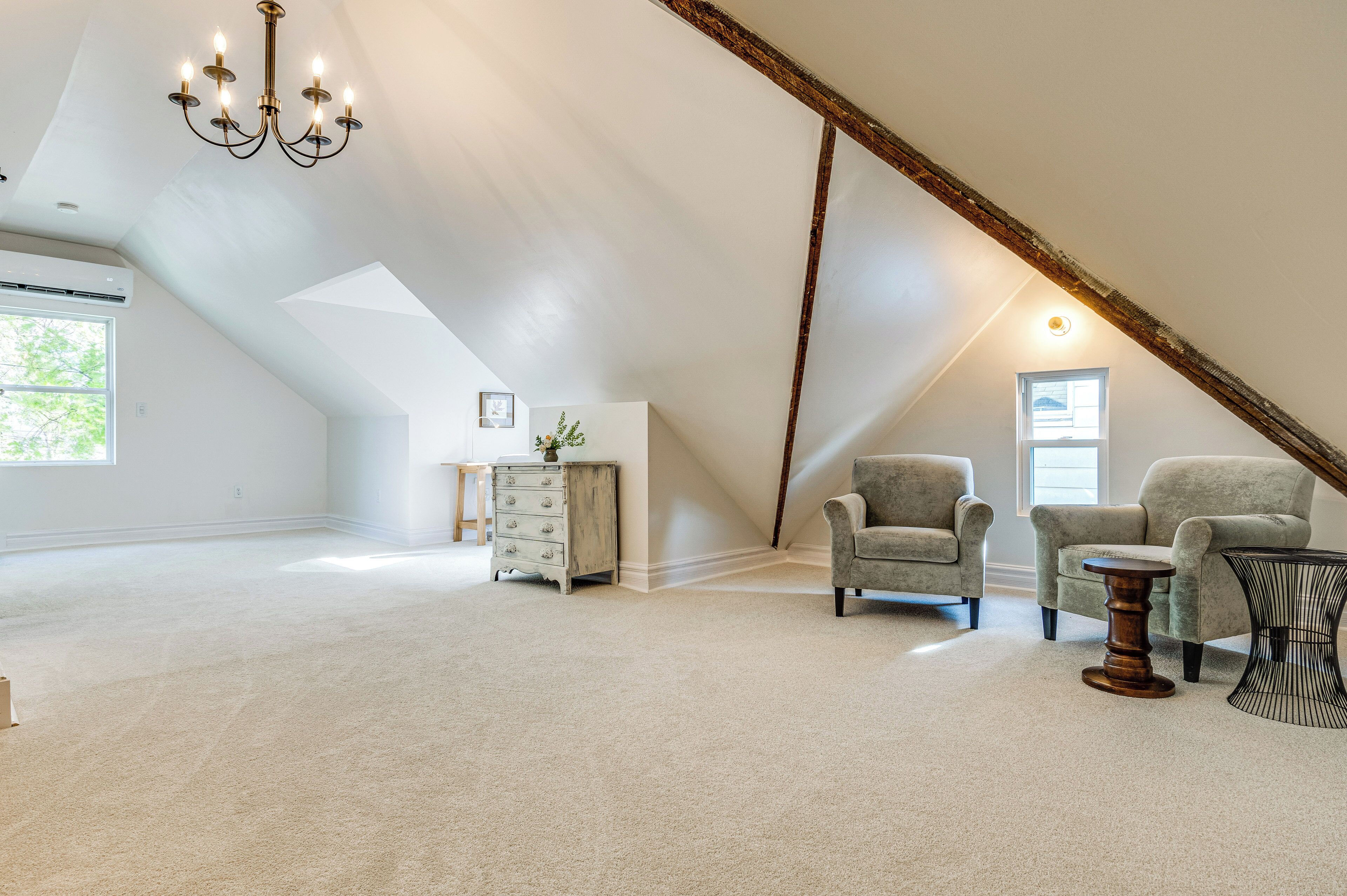 A spacious attic room with a sloped ceiling, a chandelier, two armchairs, a wooden side table, and a dresser.
