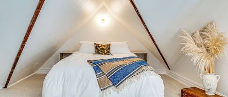 A bedroom with a sloped ceiling, a bed with white bedding, a patterned throw blanket, and a vase with pampas grass.