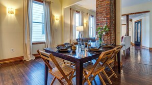A dining area with a wooden table set for six, flanked by brick walls and large windows with curtains. - Cozy Indianapolis Vacation Rental w/ Micro Farm! (Indianapolis)