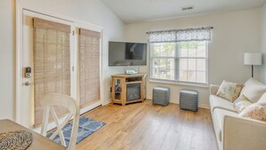 A living room with a wooden floor, a sofa, a TV on a wooden stand, a window with blinds, and a door with a lock.