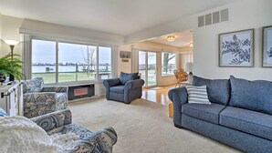 A spacious living room with a large window overlooking a body of water, a fireplace, and a cozy seating area with patterned sofas and a blue couch. - Waterfront East China Home w/ Dock & Patio! (East China)