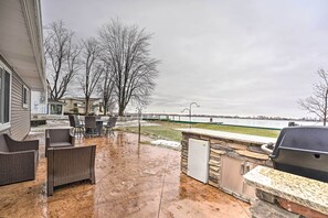 Une terrasse extérieure aménagée avec des meubles en osier, un coin barbecue en pierre et une vue sur une étendue d’eau et un pont.