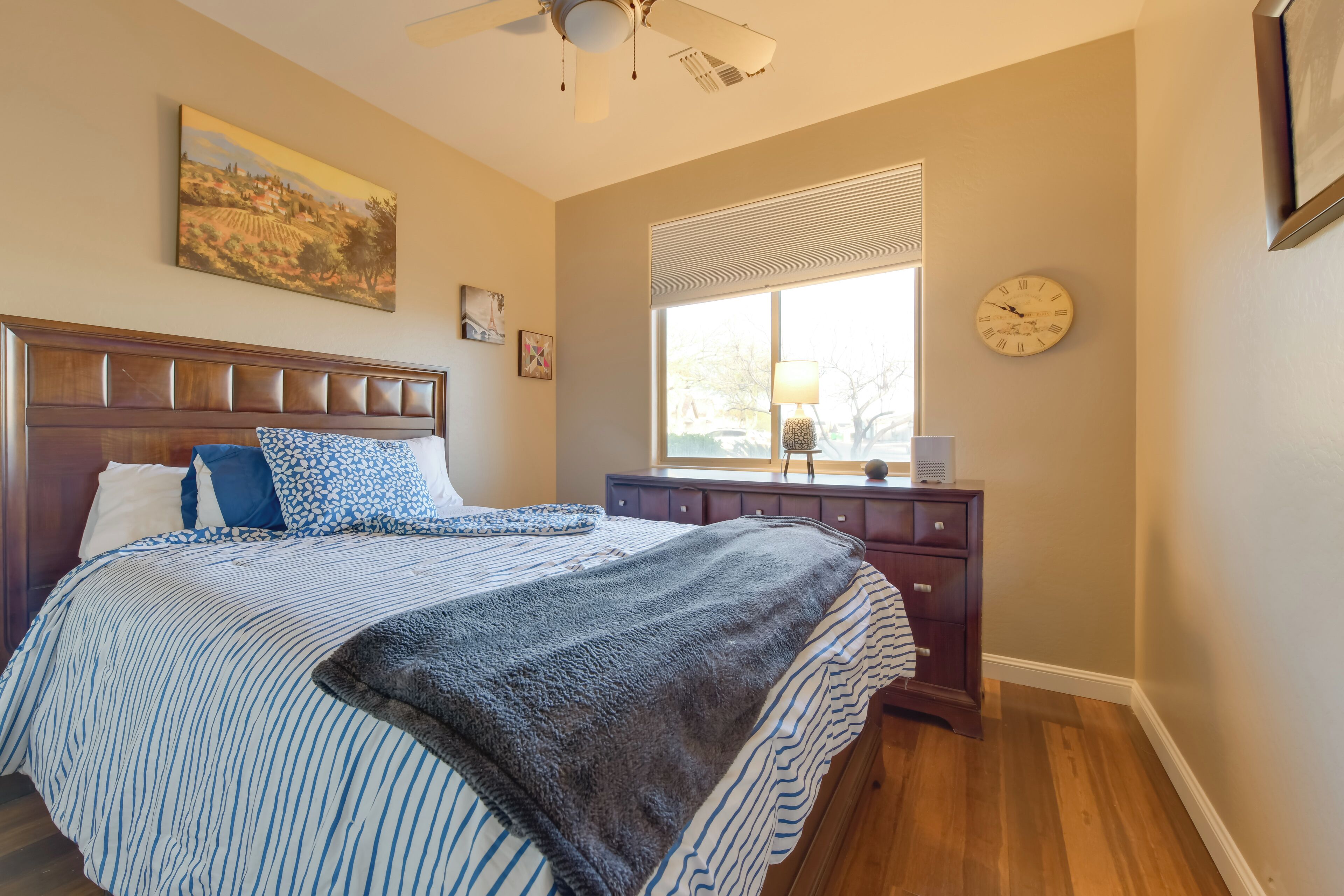 A bedroom with a large bed, a wooden headboard, a window with blinds, a clock, and a dresser.