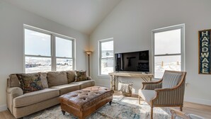 Un salon avec un canapé beige, un pouf marron, un fauteuil à rayures et un grand téléviseur à écran plat posé sur une table en bois.