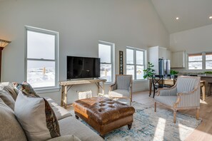 A spacious living room with a large flat-screen TV, comfortable seating, and a view of the snowy outdoors through large windows.
