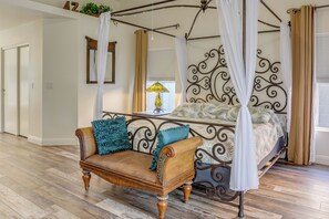 A bedroom with a four-poster bed, a sofa, a bedside table with a lamp, and a window with curtains.