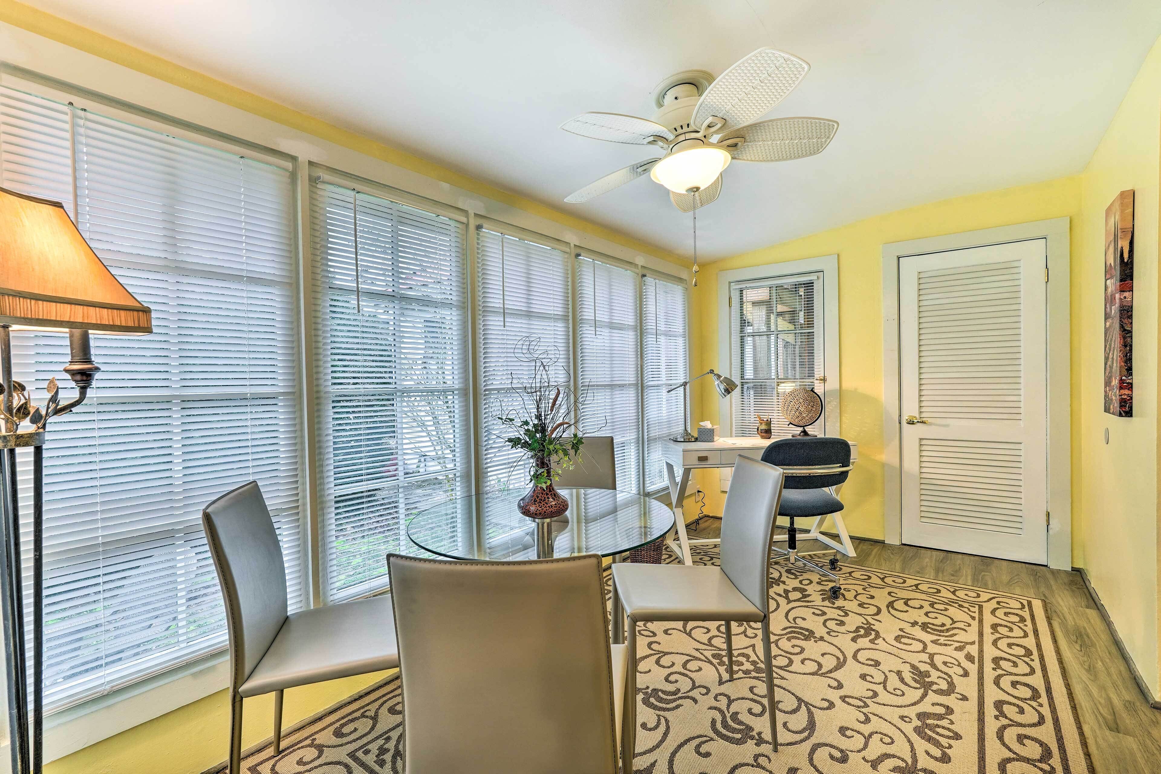 A room with a dining table, two chairs, a ceiling fan, and large windows with blinds.