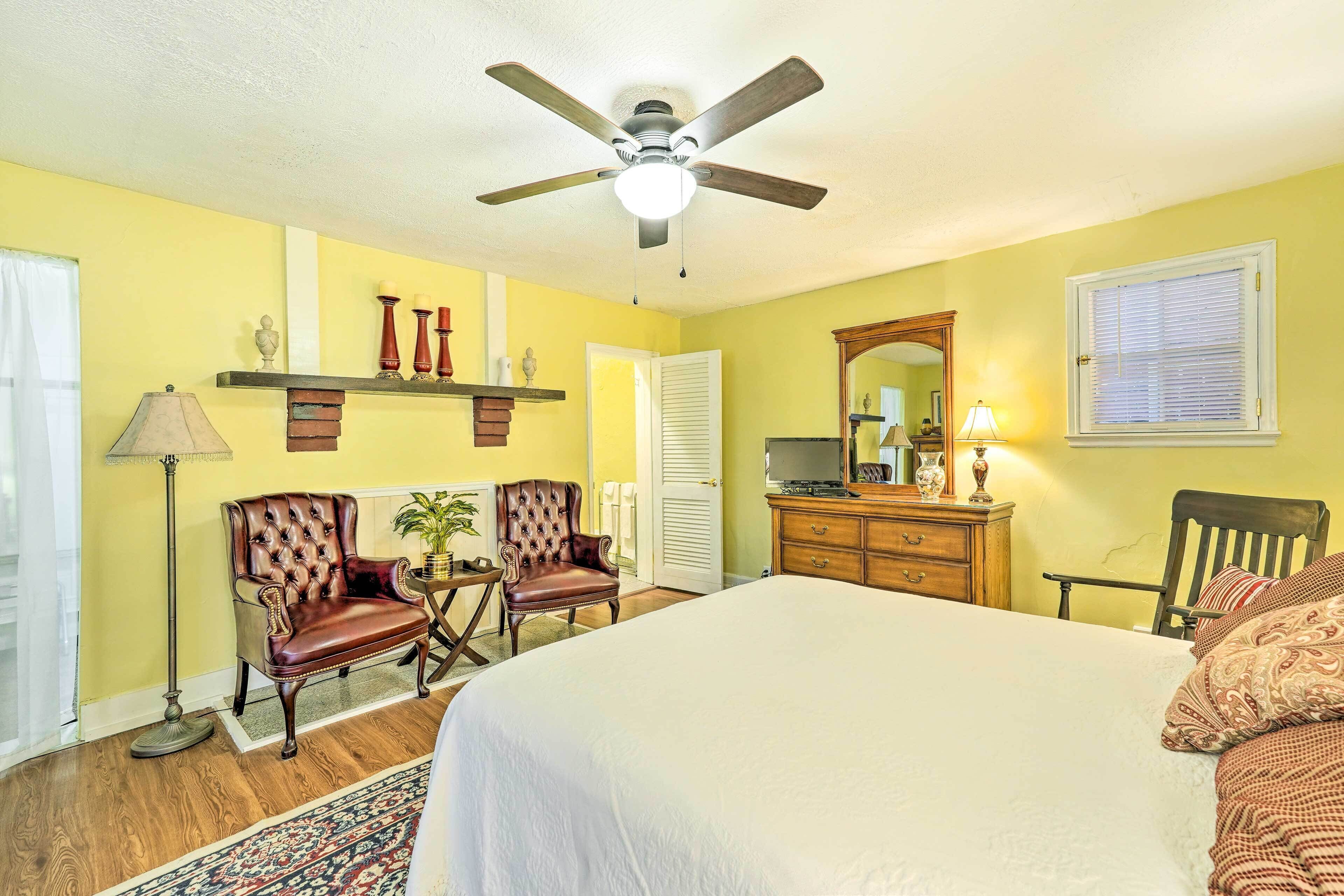 A hotel room with a bed, two armchairs, a dresser, a ceiling fan, and a window with blinds.