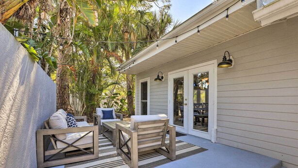 Une terrasse avec un banc en bois, une table et une chaise rembourrée. Des guirlandes lumineuses sont suspendues au-dessus, et une porte donne accès à la maison.