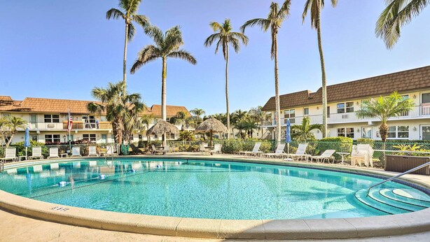 A clear swimming pool surrounded by lounge chairs and palm trees, with a building in the background. - Marco Island Condo ~ 1 Mi to the Beach! (Marco Island)