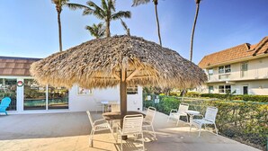 An outdoor seating area with a thatched umbrella, white metal chairs, and a pool in the background. - Marco Island Condo ~ 1 Mi to the Beach! (Marco Island)