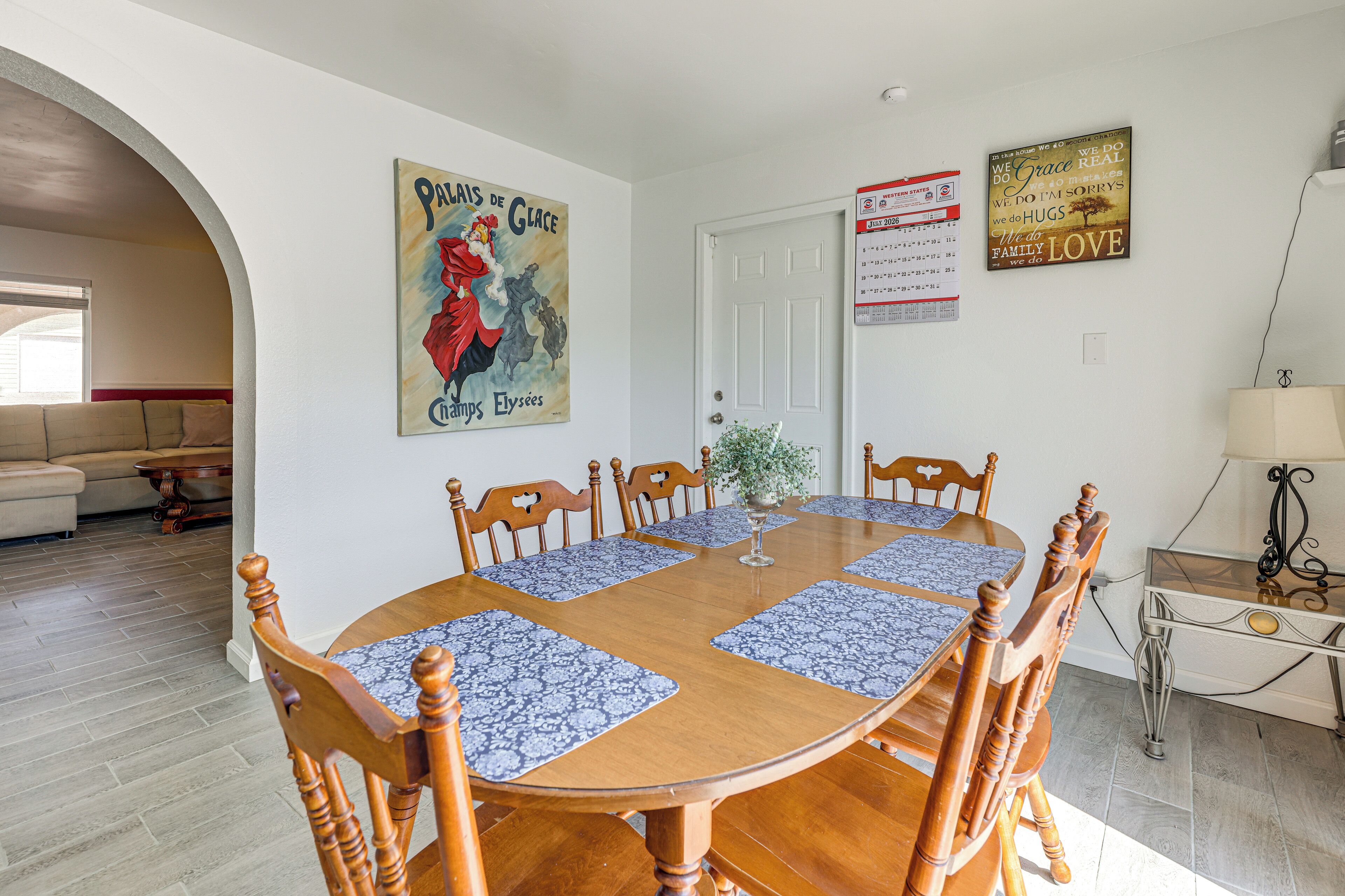Un comedor con una mesa de madera rodeada de sillas, una puerta blanca y una pared decorada con un calendario y un cartel.