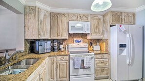 A kitchen with granite countertops, wooden cabinets, a stainless steel sink, a microwave, a stove, a refrigerator, and a hanging light fixture. - Lovely Fort Pierce Condo, Walk to Beach! (Fort Pierce)