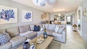 A modern living room with a large L-shaped sofa, a coffee table with decorative items, and a dining area in the background. - Lovely Fort Pierce Condo, Walk to Beach! (Fort Pierce)