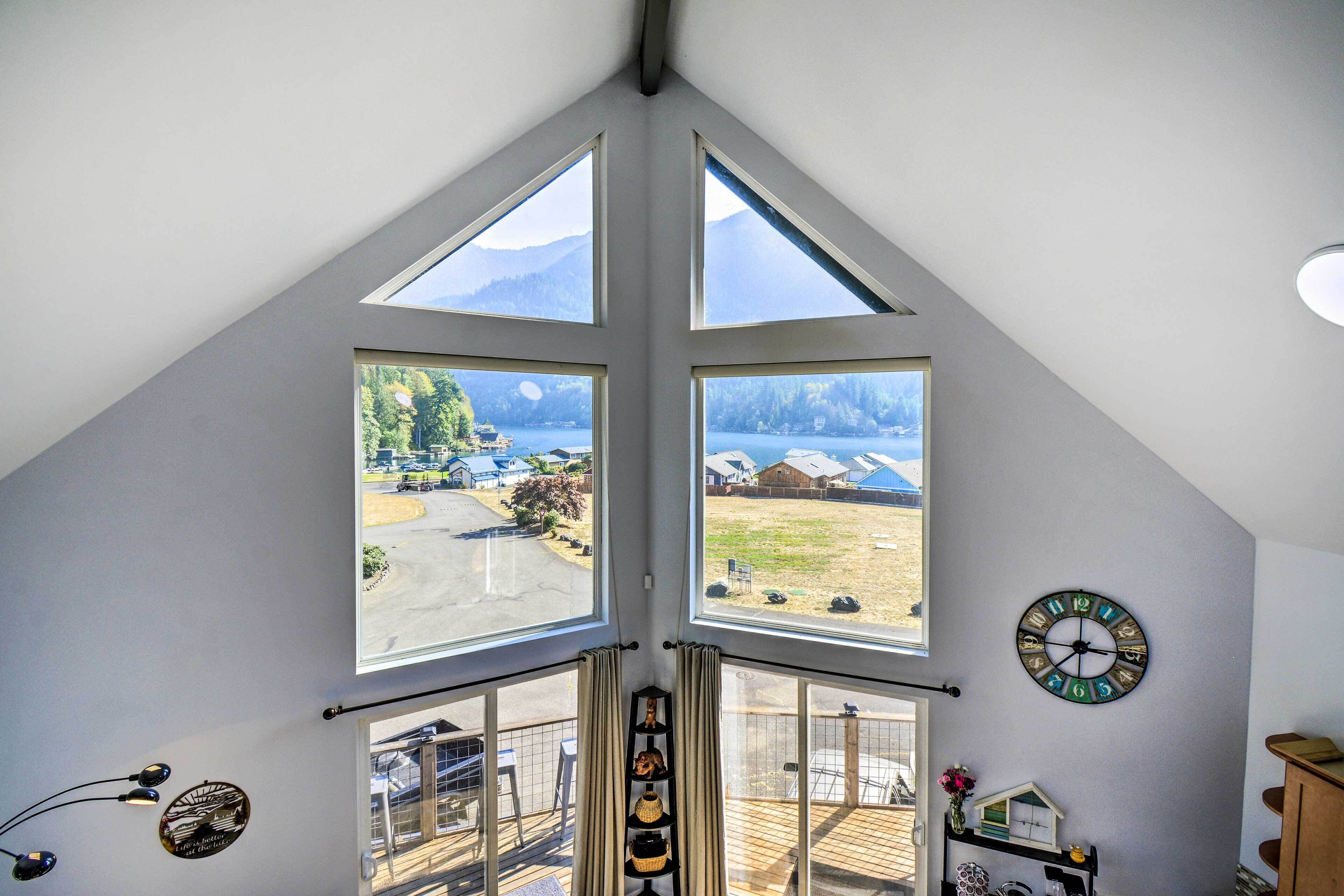 Una habitación con techo inclinado, ventanas amplias, un reloj y vistas a un lago y montañas.
