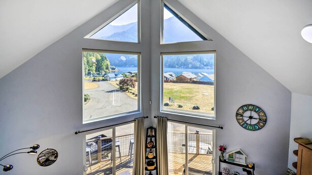 Ein Zimmer mit schrägem Dach, großen Fenstern, einer Uhr und Blick auf einen See und Berge.