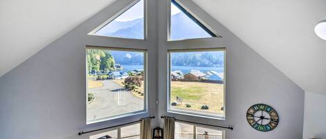 Quarto com teto inclinado, janelas amplas, um relógio e vista para um lago e montanhas.