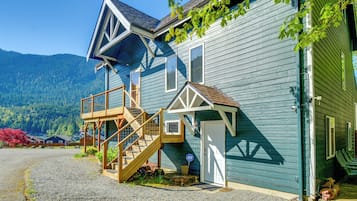 A two-story house with a wooden staircase leading to a deck, a garage door, and a mountain backdrop.