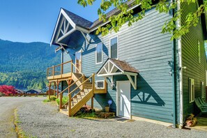 Une maison à deux étages avec un escalier en bois menant à une terrasse, une porte de garage et une vue sur les montagnes en arrière-plan.