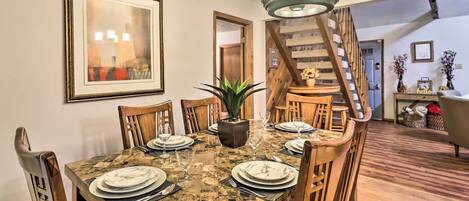 A dining area with a marble table set for four, surrounded by wooden chairs. A potted plant is on the table, and a staircase is visible in the background.